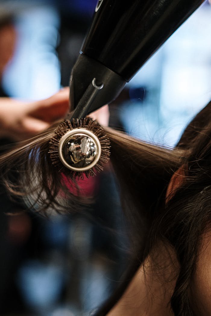 our-services-04 Close-up of a hairstylist styling hair with a brush and dryer in a salon setting.